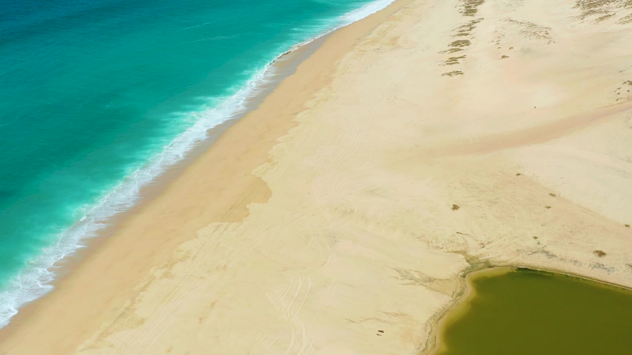 Aerial frame of La Laguna Beach Los Cabos with the shoreline running diagonally, turquoise water washing onto wet sand, dune texture across the right side, a partial green lagoon at bottom right, and no visible people.