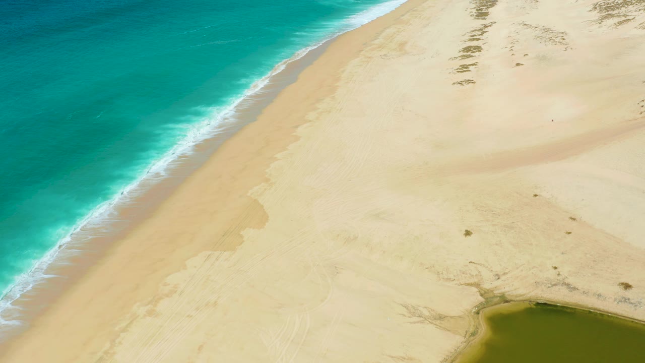 Aerial view at La Laguna Beach Los Cabos showing foamy waves along the left edge, a broad pale beach through the center, scattered dune vegetation marks near the top right, a lagoon edge at the lower right, and no visible people.