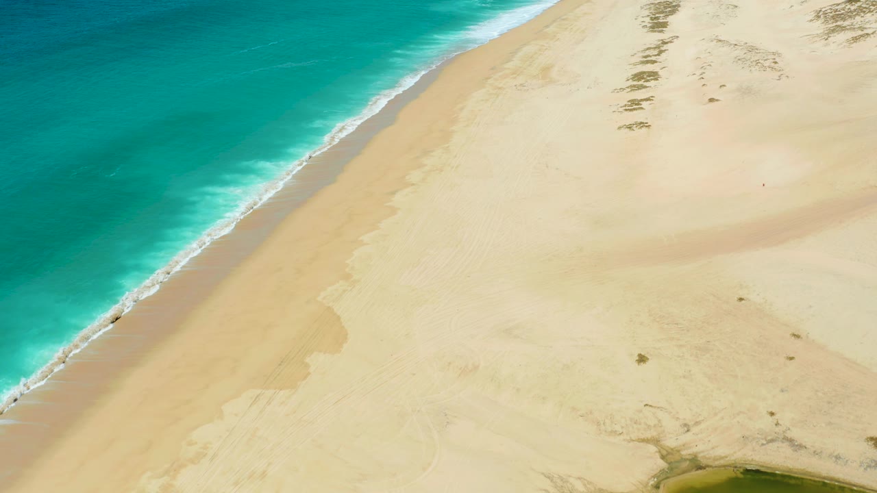 Aerial coastline with turquoise water, foam line, pale sand, and dune patterns.