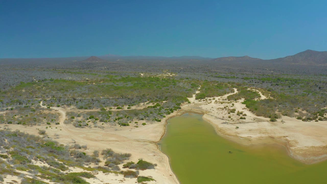 Aerial frame at La Laguna Beach Los Cabos with the green waterhole lower in view, sandy flats and brush in the midground, mountains on the horizon, and no visible people.