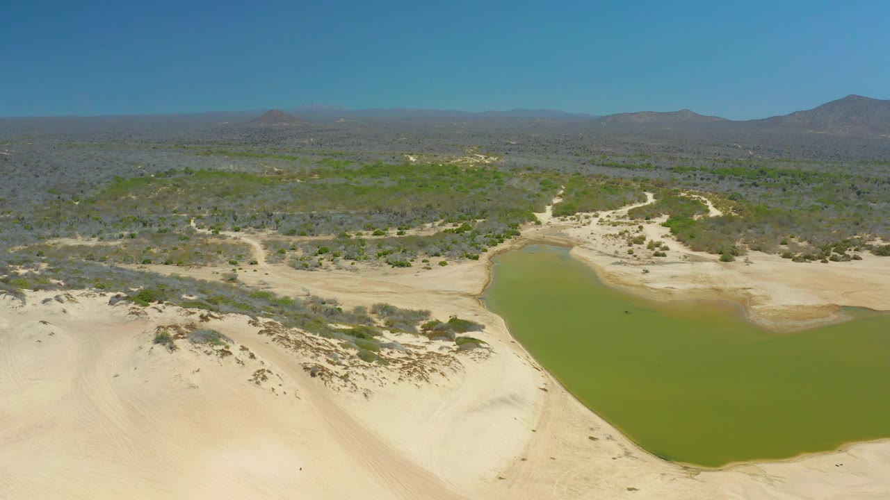 Wide aerial frame from La Laguna Beach Los Cabos showing the lagoon along the lower right, pale dune ridges across the foreground, scrubland in the distance, mountains on the horizon, and no visible people.
