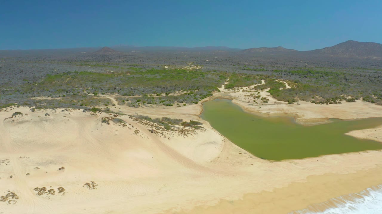 At 0:01, the La Laguna Beach Los Cabos lagoon sits centered behind broad sand dunes, with scrubland and mountains filling the horizon and no visible people.