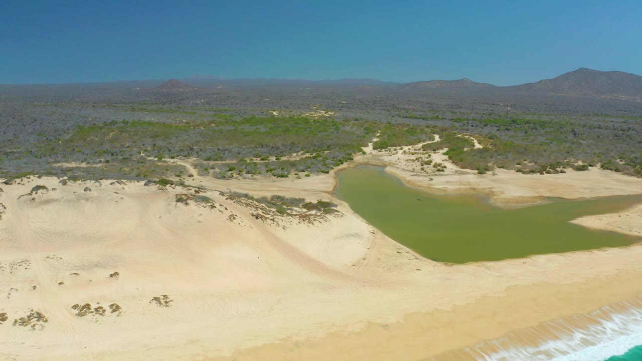 At La Laguna Beach Los Cabos, the lagoon extends across the midground while pale dunes and low desert vegetation lead toward distant mountains, with no visible people.