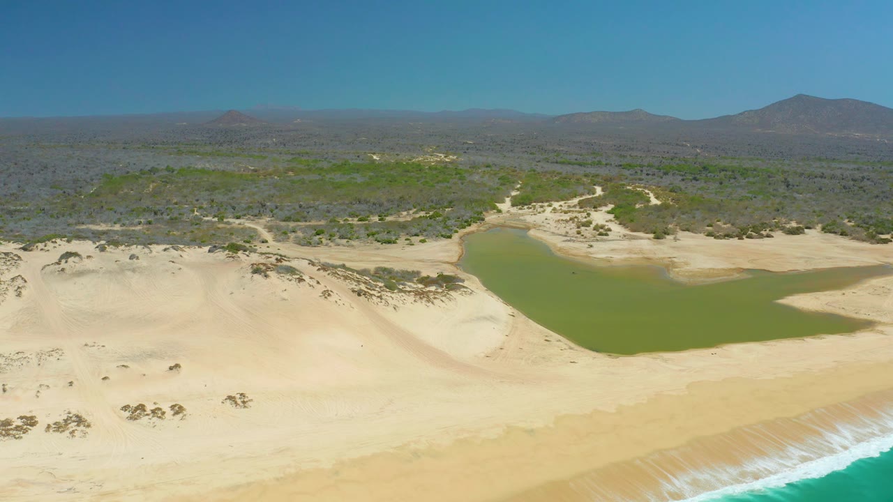 At 0:03, a turquoise wave line appears along the lower right foreground while the La Laguna Beach Los Cabos lagoon and dunes remain steady behind it, with no visible people.