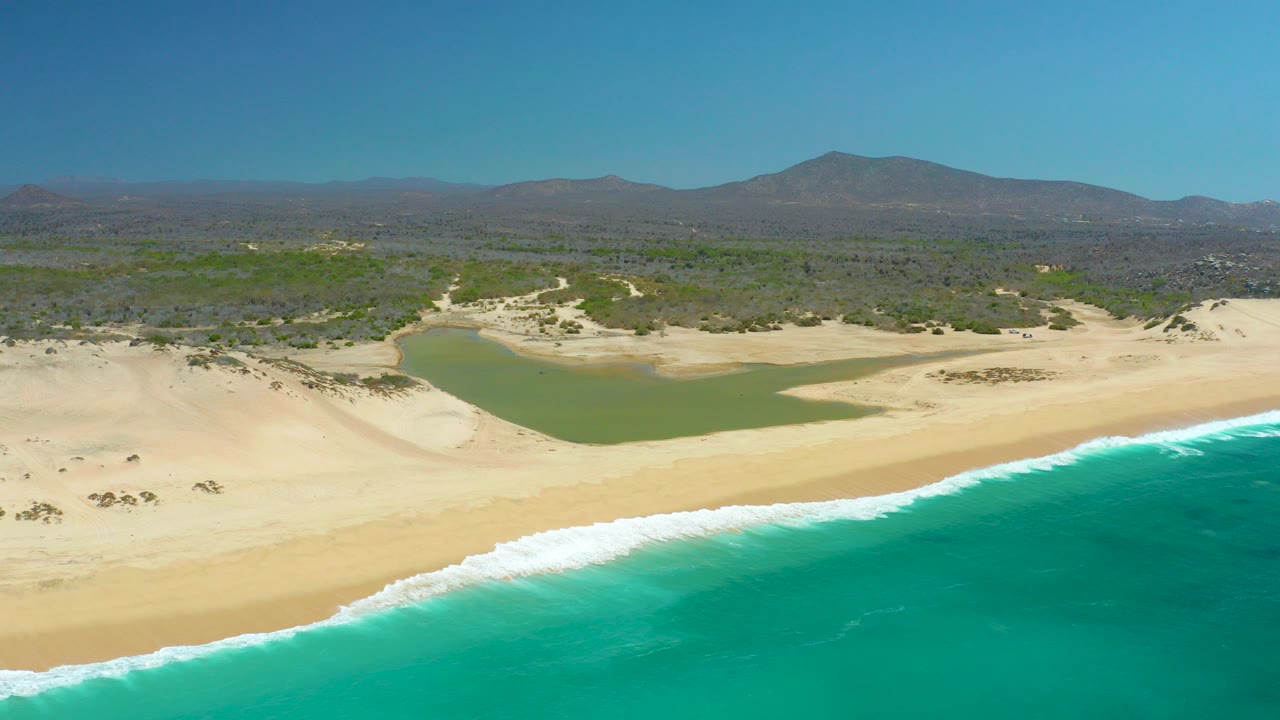 At La Laguna Beach Los Cabos, turquoise ocean and white foam meet a wide sandy shore below a green lagoon, dunes, scrubland, distant mountains, and clear sky, with no visible people.