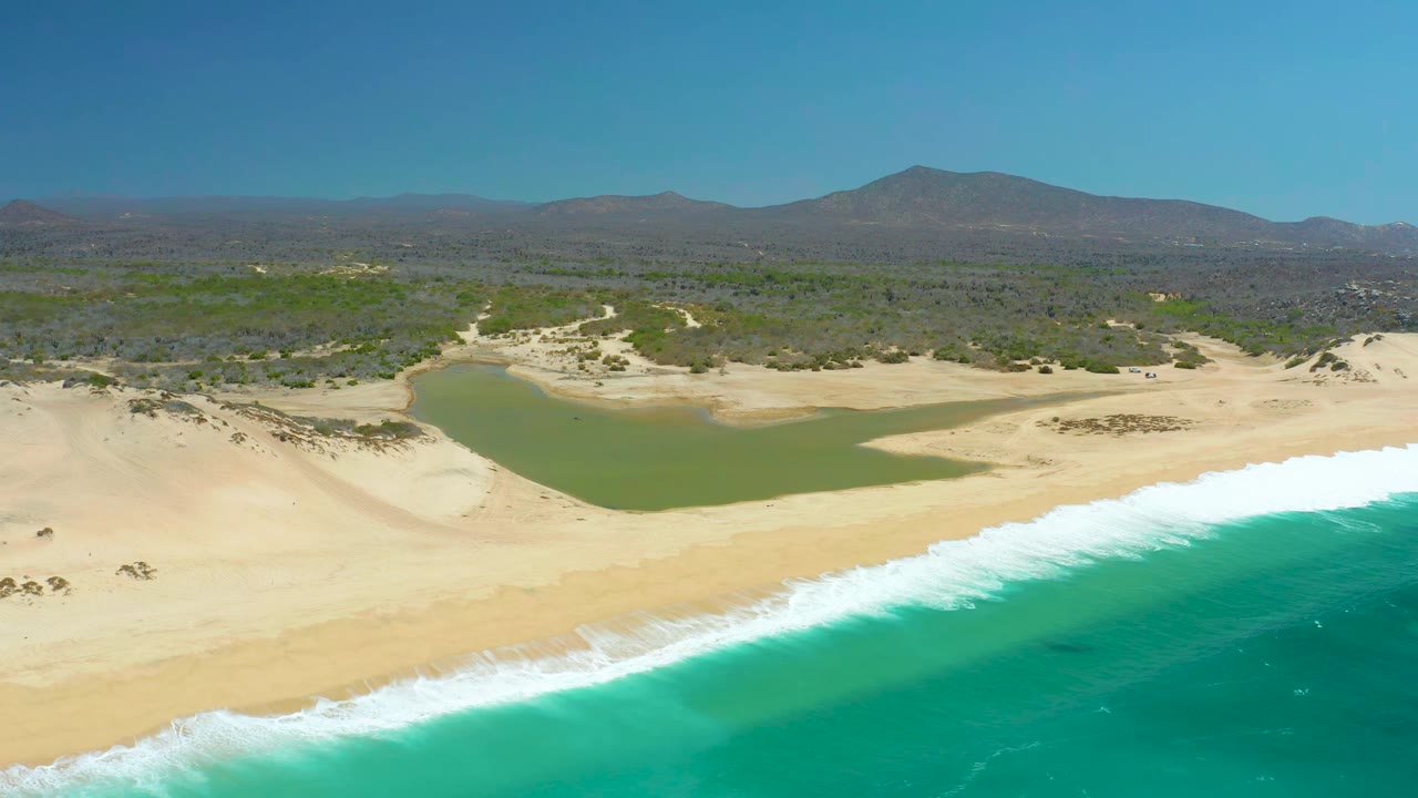 Aerial view at La Laguna Beach Los Cabos, Mexico, with the breaking white surf slightly farther along the lower beach, the lagoon centered behind the sand, and no visible people.