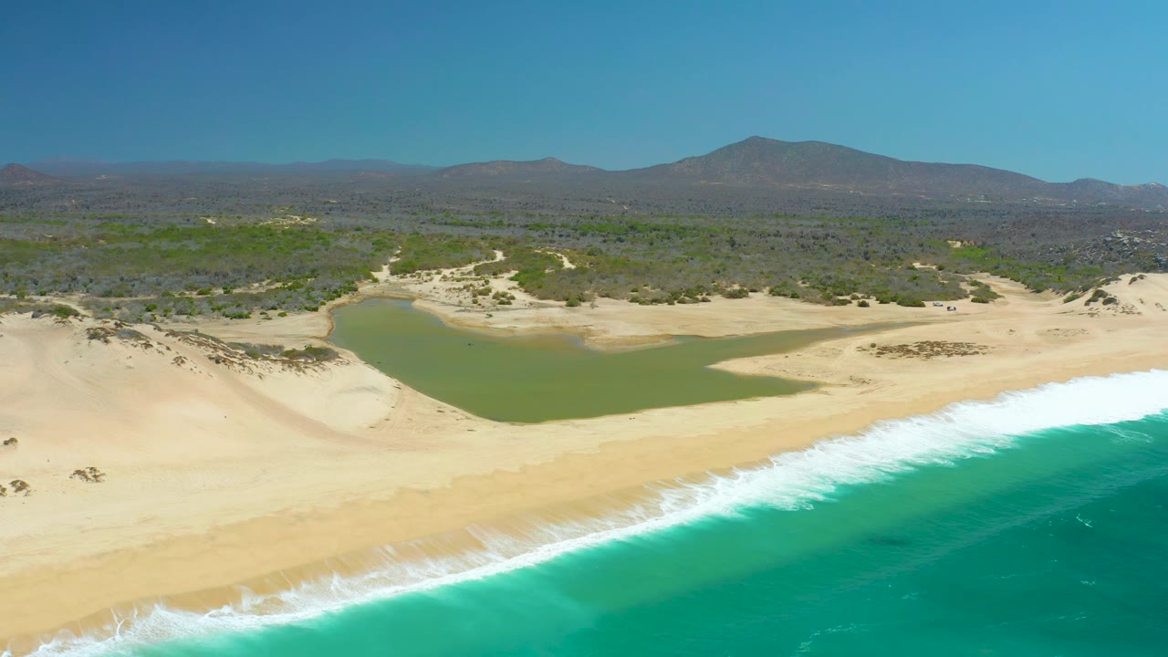 Aerial frame from La Laguna Beach Los Cabos, Mexico, where the turquoise ocean fills more of the lower right, white surf runs diagonally across the sand, and no visible people appear.