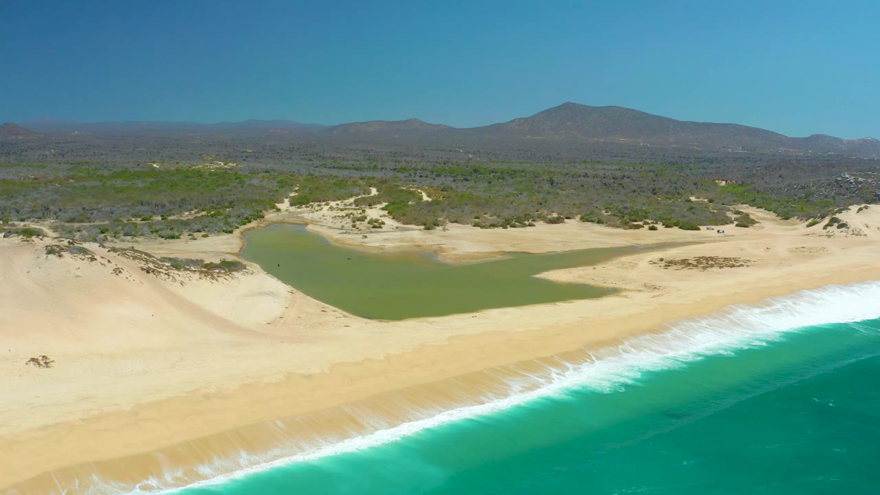 Mid-clip aerial view over La Laguna Beach Los Cabos, Mexico, with the lagoon shifted left of center, surf cutting across the lower right, and no visible people on the open sand.
