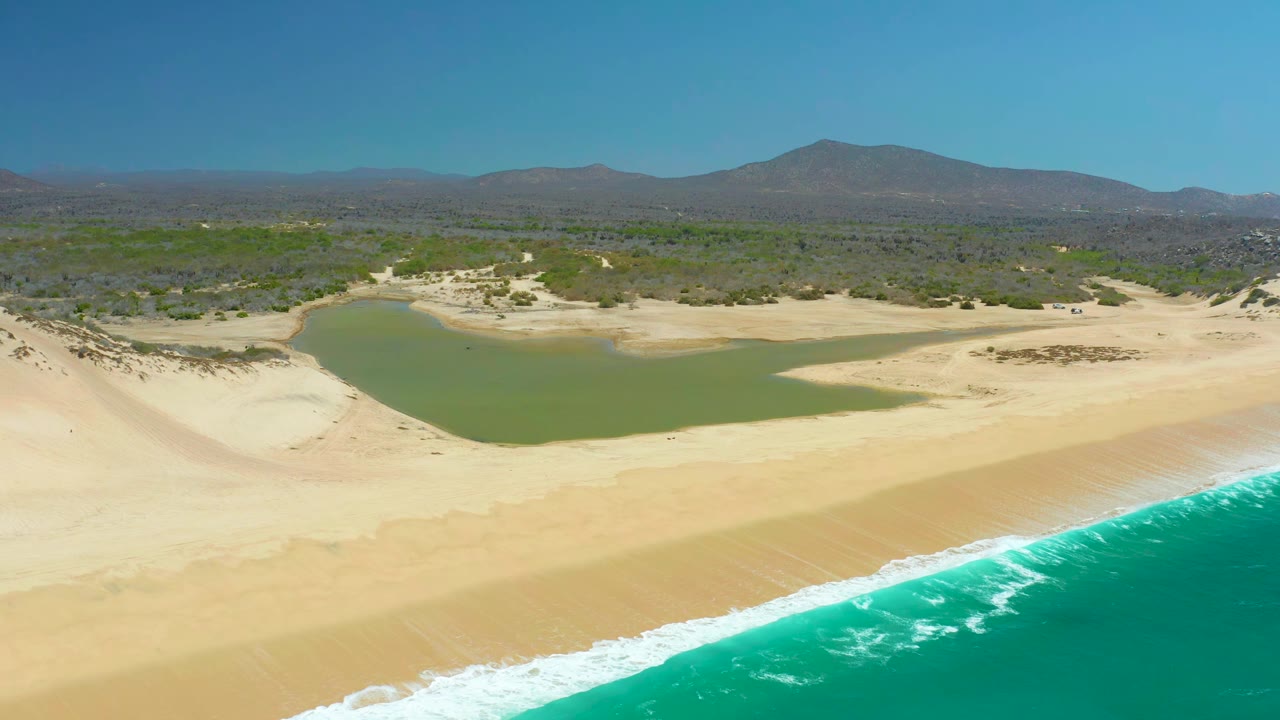 Wide aerial view at La Laguna Beach Los Cabos with white wave foam edging the sand, the green lagoon centered inland, low scrub beyond it, distant mountains, blue sky, and no visible people.