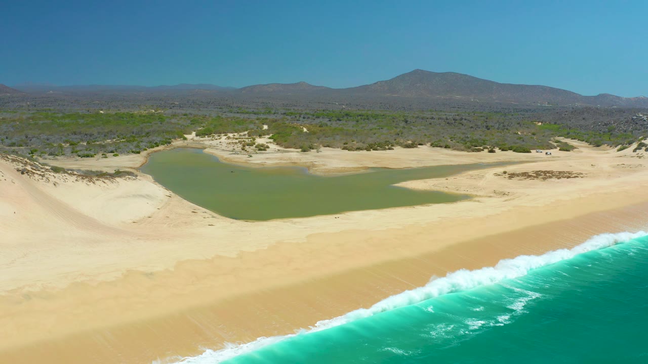 Aerial shoreline frame from La Laguna Beach Los Cabos showing a fuller band of breaking white surf in front, broad tan sand, a green lagoon behind the dunes, scrubland, mountain horizon, and no visible people.