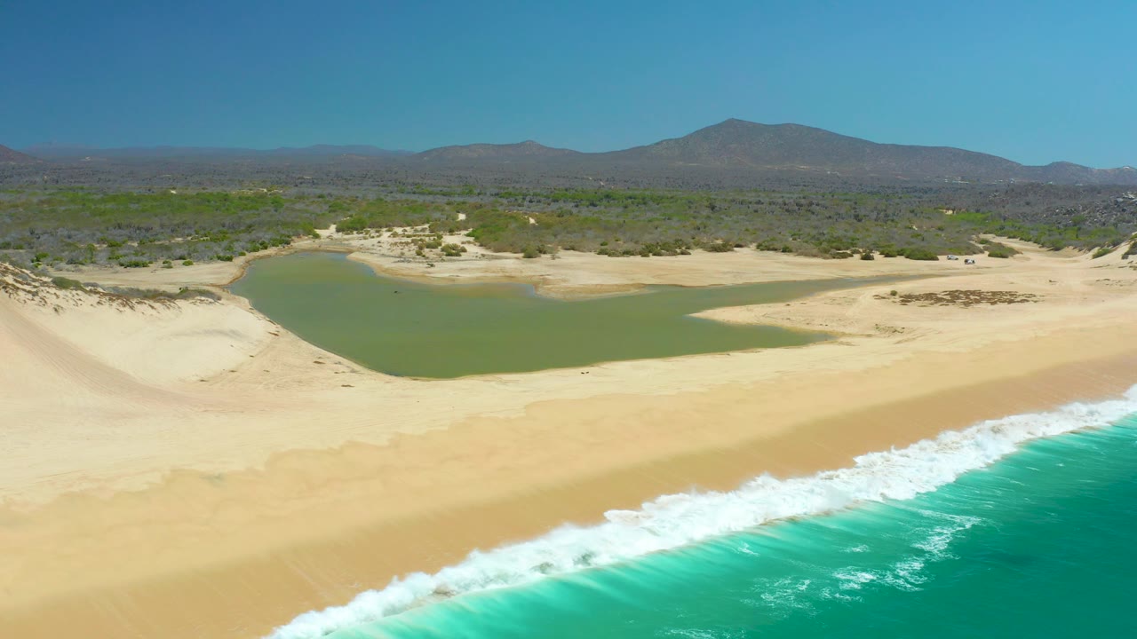 Drone-height view of La Laguna Beach Los Cabos with ocean foam crossing the foreground, a smooth sandy beach, the green lagoon just inland, scattered vegetation, hazy distant mountains, and no visible people.