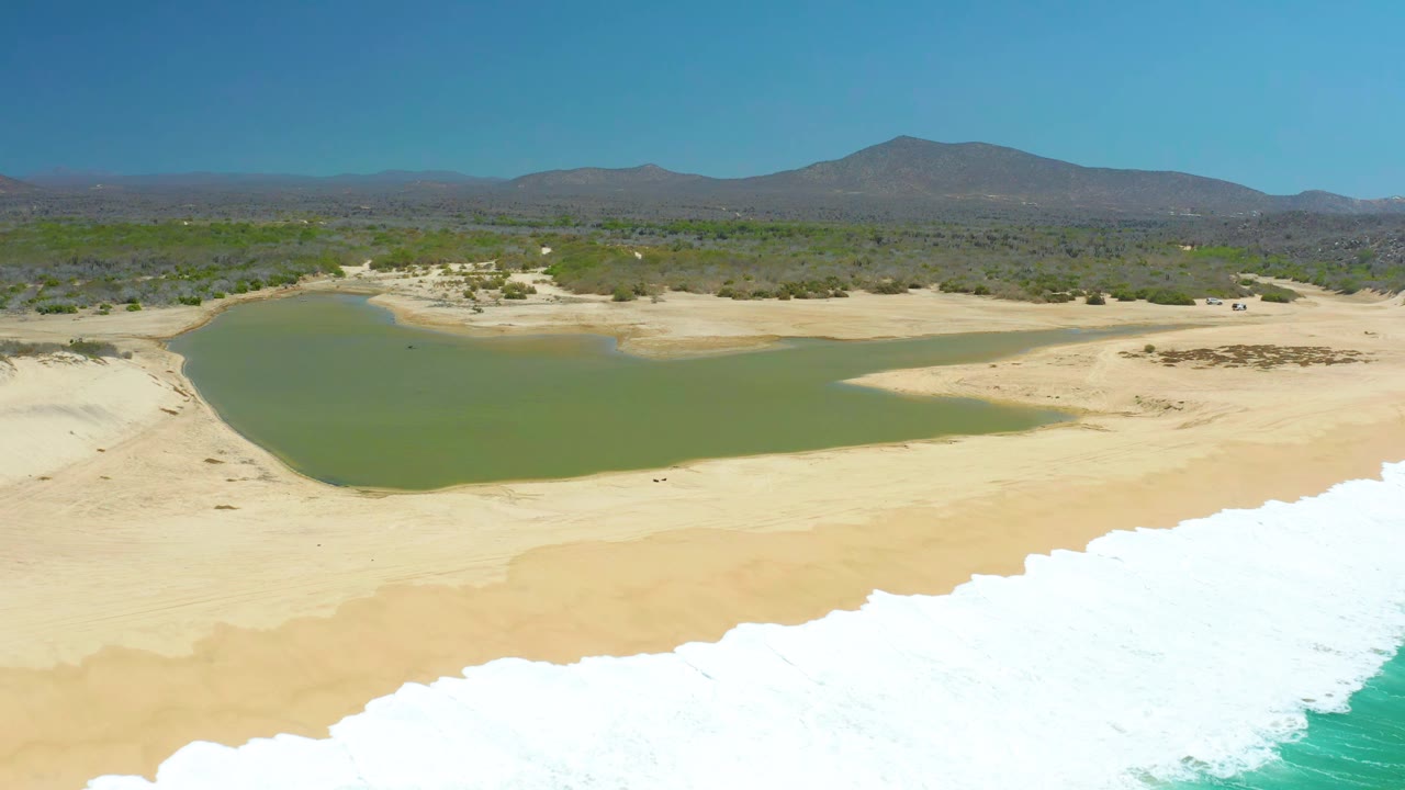 At La Laguna Beach Los Cabos, the aerial view holds on the green lagoon and sandy shore while a broad white surf band fills the lower edge, with no visible people.