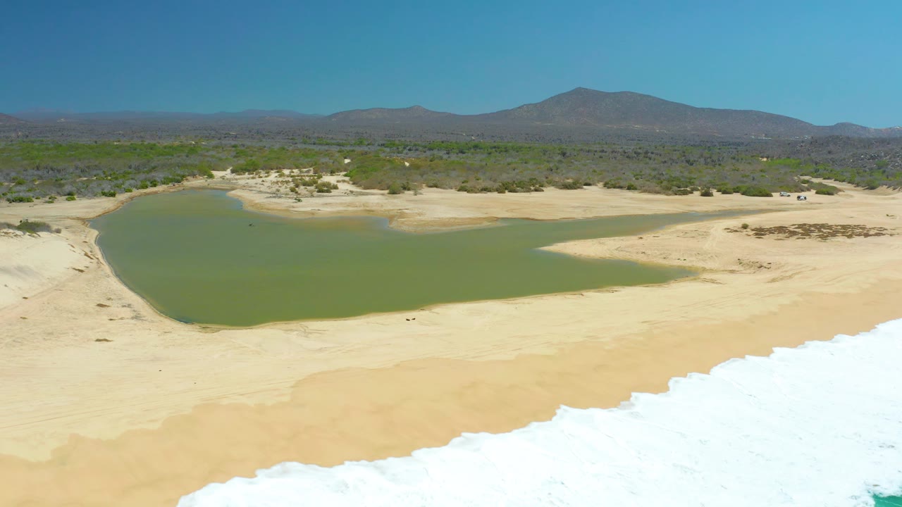 At La Laguna Beach Los Cabos, the lagoon appears slightly farther right in frame with pale sandbanks around it, white surf foam below, and no visible people.