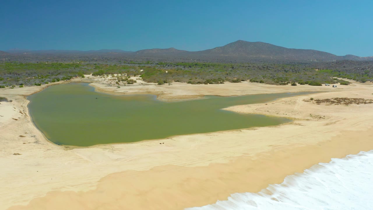 At La Laguna Beach Los Cabos, the aerial frame shows more of the lagoon's left shoreline, pale sand stretching toward the surf, and no visible people.