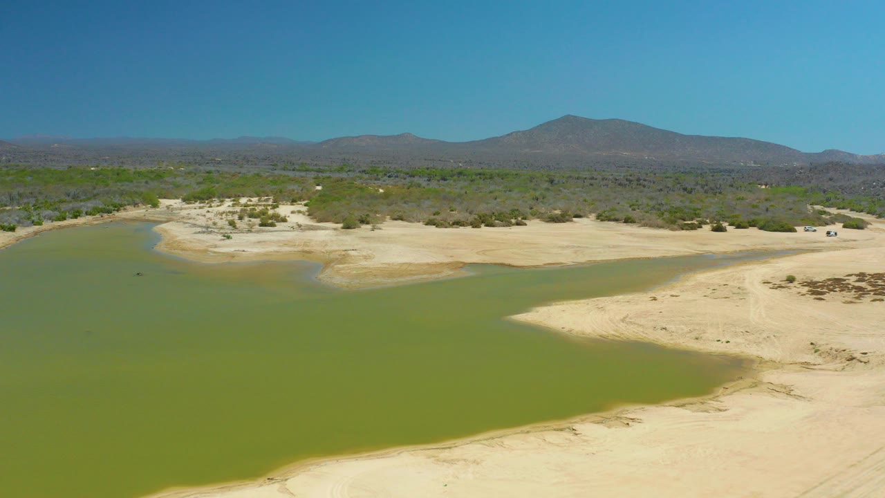 Green lagoon water beside pale sandy riverbed with desert vegetation and mountains.