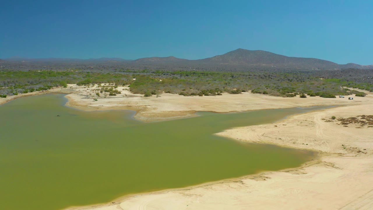 Aerial frame at La Laguna Beach Los Cabos with green water curving around exposed sandbars, low desert vegetation, mountain ridges, blue sky, and no visible people.