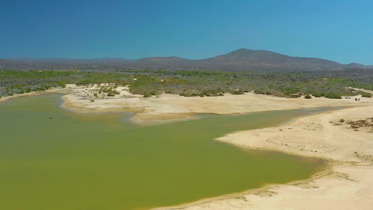 La Laguna Beach Los Cabos aerial view with green lagoon water filling the foreground, pale dry banks at right and center, sparse vegetation, distant mountains, and no visible people.