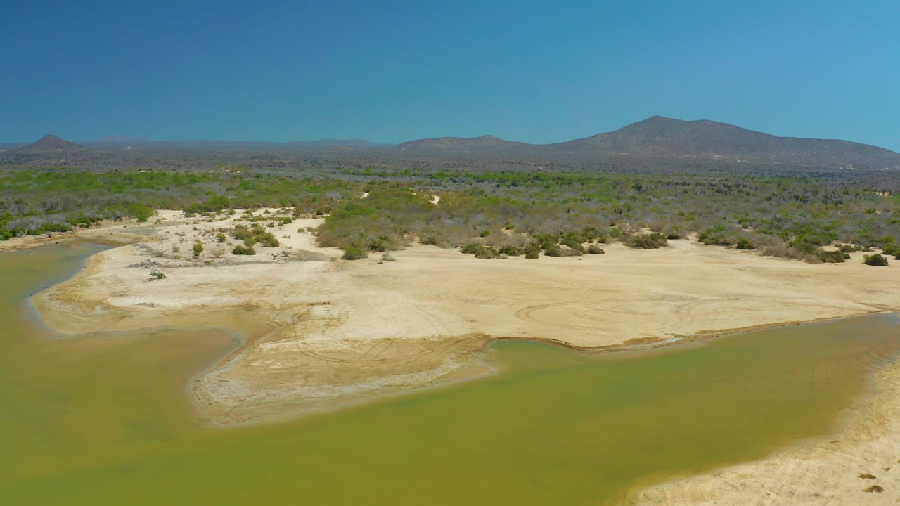 Drone view over La Laguna Beach Los Cabos showing a pale sandbar bordered by green water, patchy desert brush, low mountains in the distance, a clear sky, and no visible people.