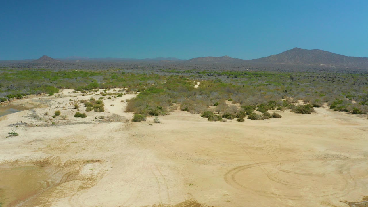 Wide aerial frame over La Laguna Beach Los Cabos sandy terrain, with tire marks in the foreground, sparse shrubs beyond, distant mountains under a clear blue sky, and no visible people.