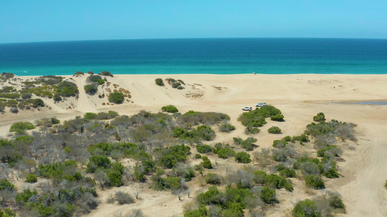 Aerial view at La Laguna Beach Los Cabos with the white SUV slightly farther left on the sand, low green shrubs below, and layered blue ocean across the horizon, with no visible people.