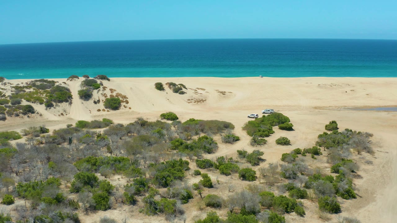 Aerial frame from La Laguna Beach Los Cabos showing the white SUV on a sandy track between scrub patches, with empty beach and turquoise water in the background, with no visible people.