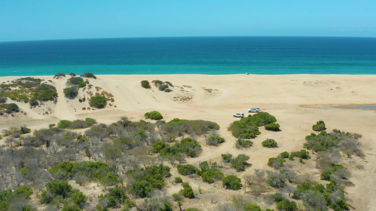 Aerial view over La Laguna Beach Los Cabos with the white SUV farther across the frame, pale dune sand, dry brush, green shrubs, and a calm blue ocean band, with no visible people.