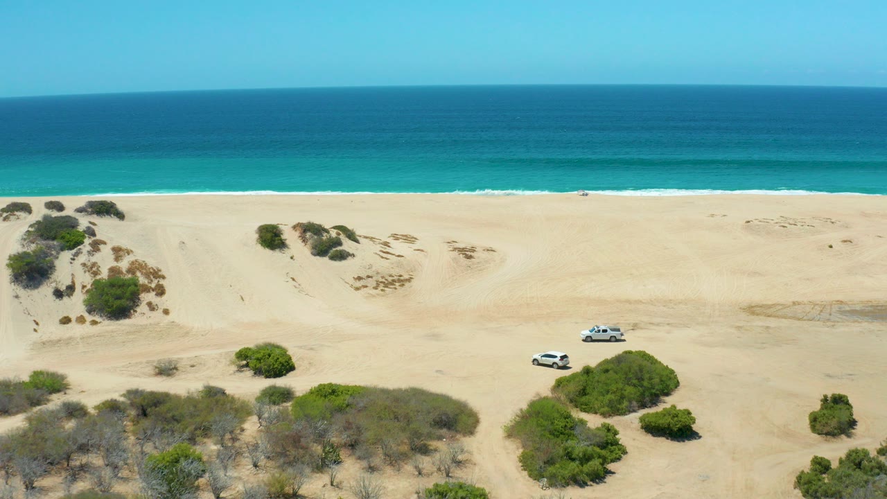 At La Laguna Beach Los Cabos, a white SUV sits high in the sandy dune area with green bushes below, turquoise surf behind, clear sky above, and no visible people.
