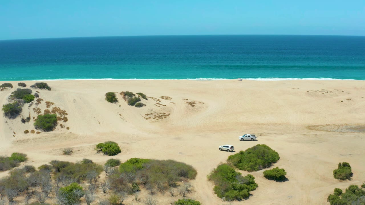 At La Laguna Beach Los Cabos, the white SUV has progressed across open sand near scattered shrubs, with a turquoise water band and dark blue horizon in the background and no visible people.
