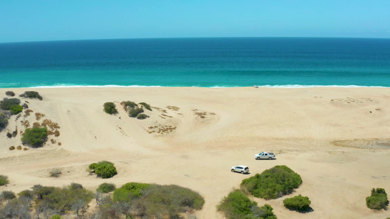 At La Laguna Beach Los Cabos, the SUV is farther across the sandy midground near rounded green bushes, with small white waves marking the shoreline and no visible people.