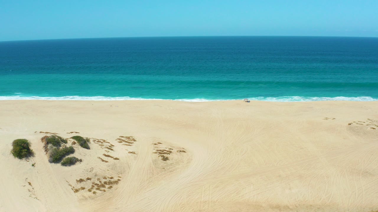 Opening aerial frame of La Laguna Beach Los Cabos with pale sand, turquoise shore break, darker blue ocean, dune vegetation near the lower left, and no visible people.