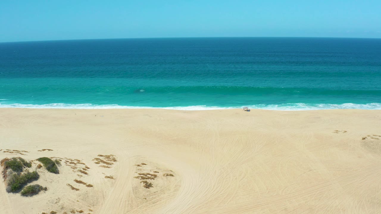 Beach and ocean aerial with dune vegetation entering from the lower left and surf across the shore.