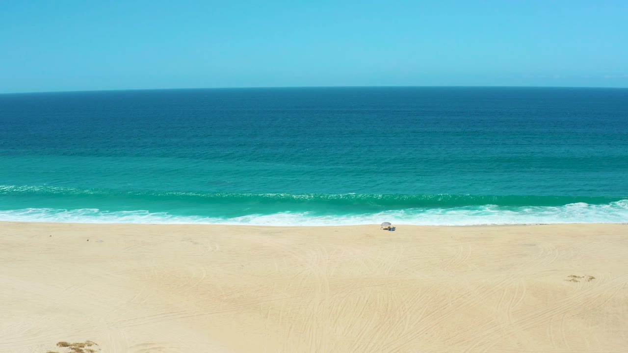 A light band of surf breaks across the La Laguna Beach shoreline, with turquoise water between the white foam and darker blue ocean, and no visible people.