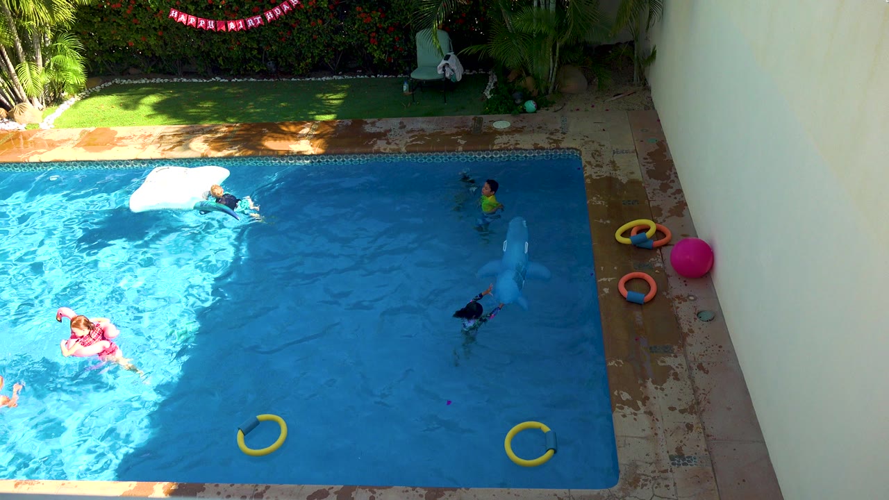 High-angle view in San Jose Del Cabo, Baja California Sur, with children visible in a backyard pool, yellow rings in the water, a pink ball on the deck, and a birthday banner above the greenery.