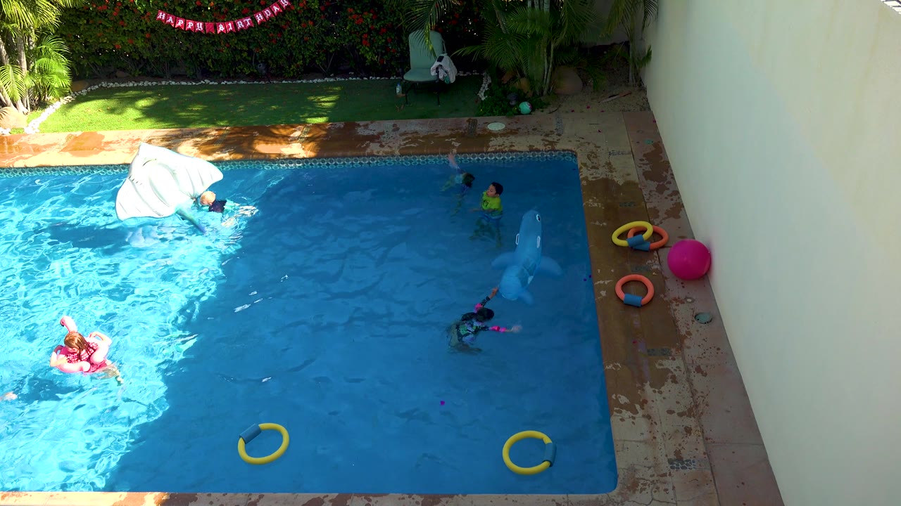 A white inflatable float turns near the upper left of the blue pool while children are visible around the water, with yellow rings below and a bright pink ball on the right-side deck.
