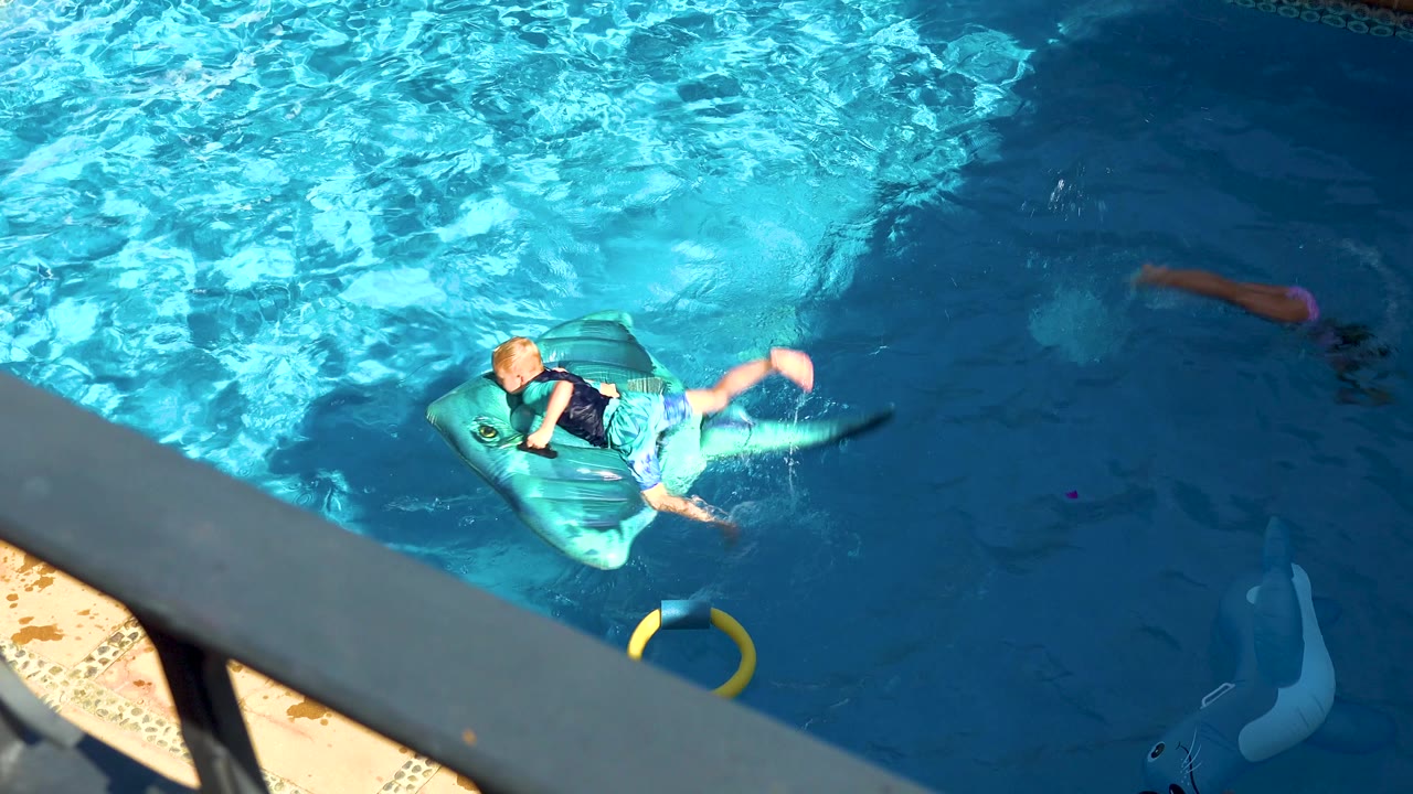 High-angle frame in San Jose Del Cabo shows a child lying on a turquoise inflatable float while other children swim nearby in blue pool water.