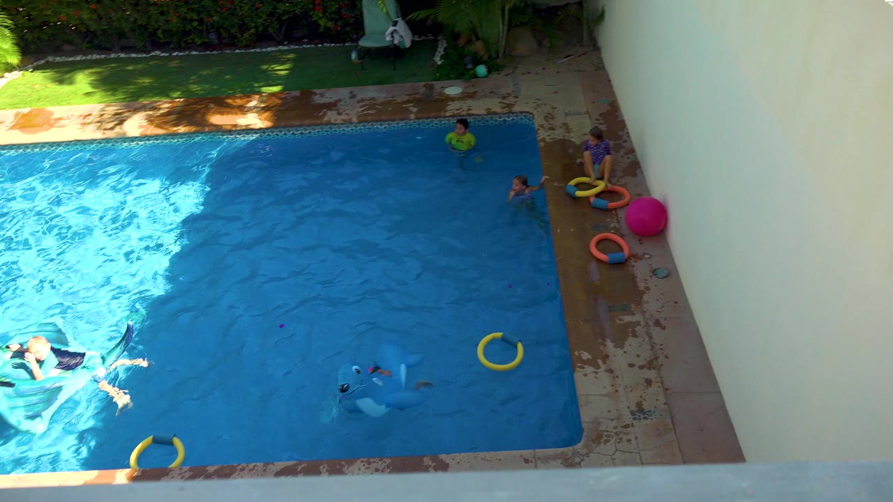 Overhead frame in San Jose Del Cabo, Mexico showing children in a blue backyard pool, a yellow ring in the water, pool toys on the wet deck, greenery, and a pale wall.