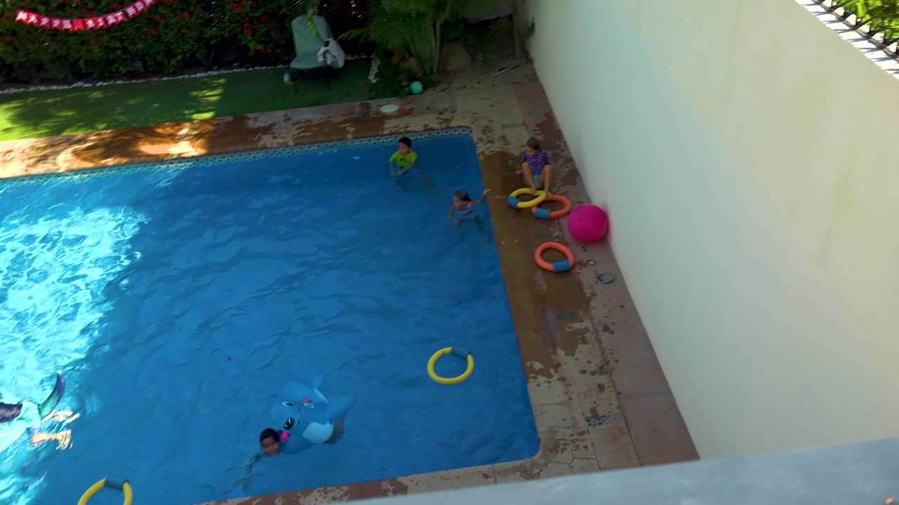 Overhead view in San Jose Del Cabo, Mexico as children move through the pool near a blue shark-shaped float, with a yellow ring in the water and more toys along the wet deck.