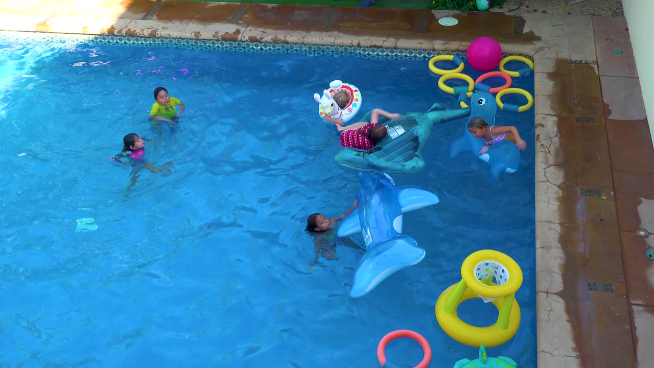 Children in San Jose Del Cabo play across a blue backyard pool while a child leans on a dark inflatable dolphin float and another holds a large blue dolphin float near the center.