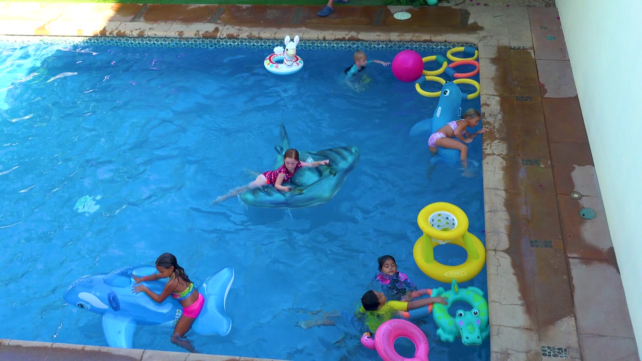 Children play in a blue swimming pool in San Jose Del Cabo, Mexico, with a girl on a shark float, another child near a blue dolphin float, and colorful rings along the pool edge.