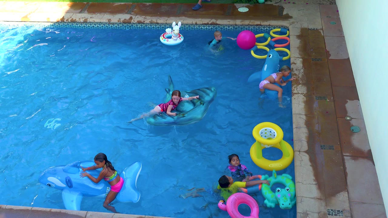 Children continue playing in the San Jose Del Cabo pool as the shark float sits near center, a child holds the blue float in the foreground, and yellow rings line the right side.