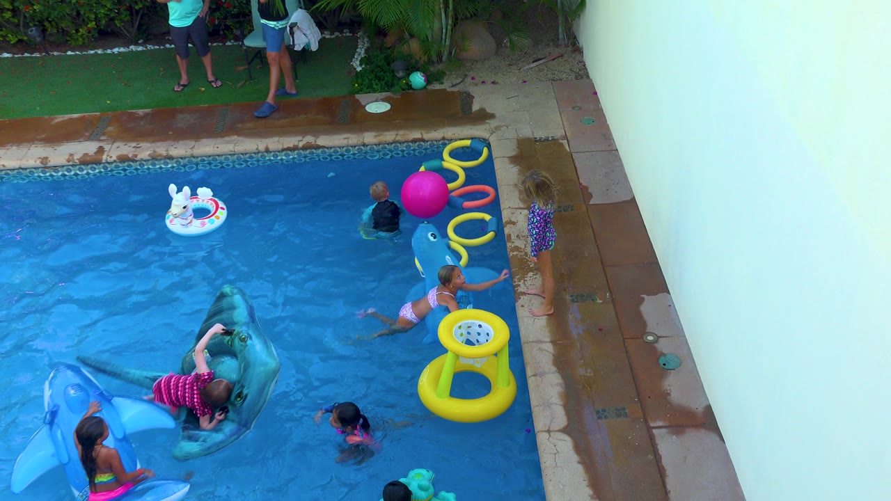 Children in San Jose Del Cabo play in a backyard swimming pool with a blue whale float, yellow rings, a pink ball, and a rabbit ring visible around the water.