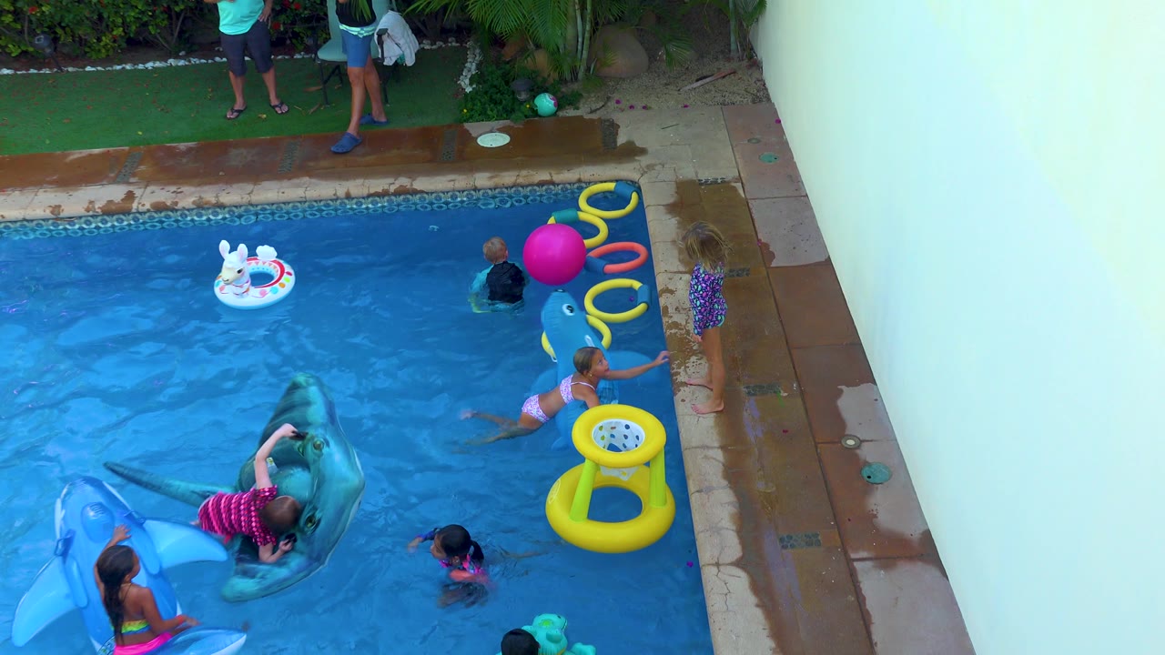 Children continue playing in the San Jose Del Cabo backyard pool as two adults are partly visible near the turf and a child holds near a yellow inflatable ring.