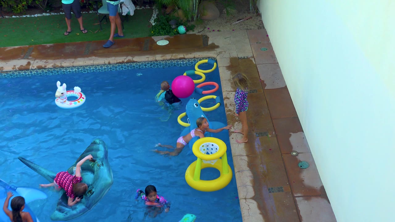 A child lies across the blue whale float while other children swim nearby in the San Jose Del Cabo backyard pool, with yellow rings and a pink ball near the deck.