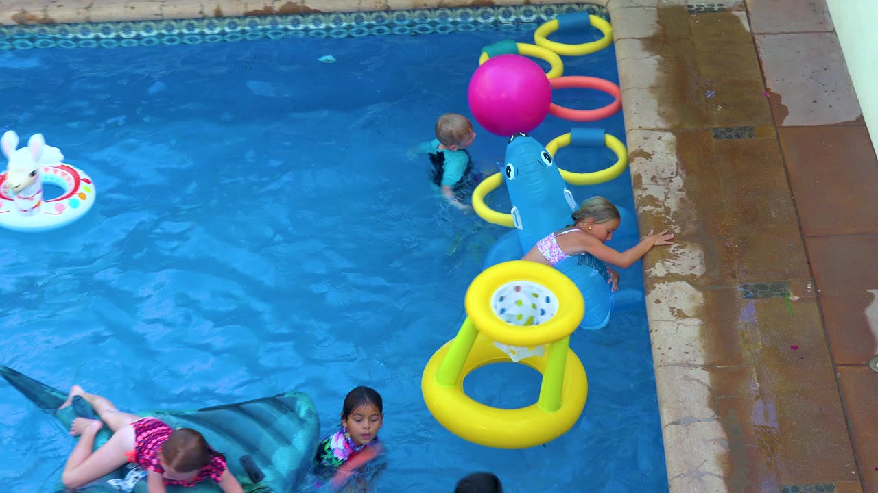 Children play in a San Jose Del Cabo pool with a blue shark float near the deck, a yellow ring float, a pink ball, and other colorful inflatable toys.