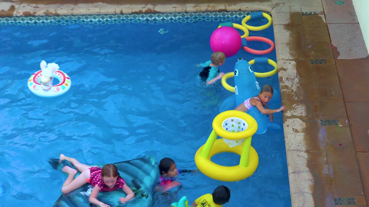 A child rests on a dark green float while another child stays beside the blue shark float near the San Jose Del Cabo pool deck and colorful rings.