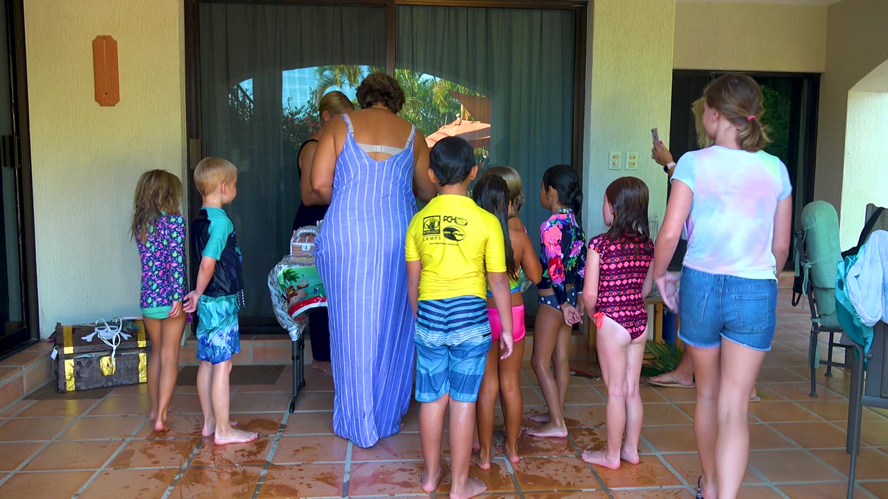 Children in swimwear stand in a line-like cluster on the San Jose del Cabo patio while adults gather around them, with a green chair and light-colored towel or bag at the right edge.