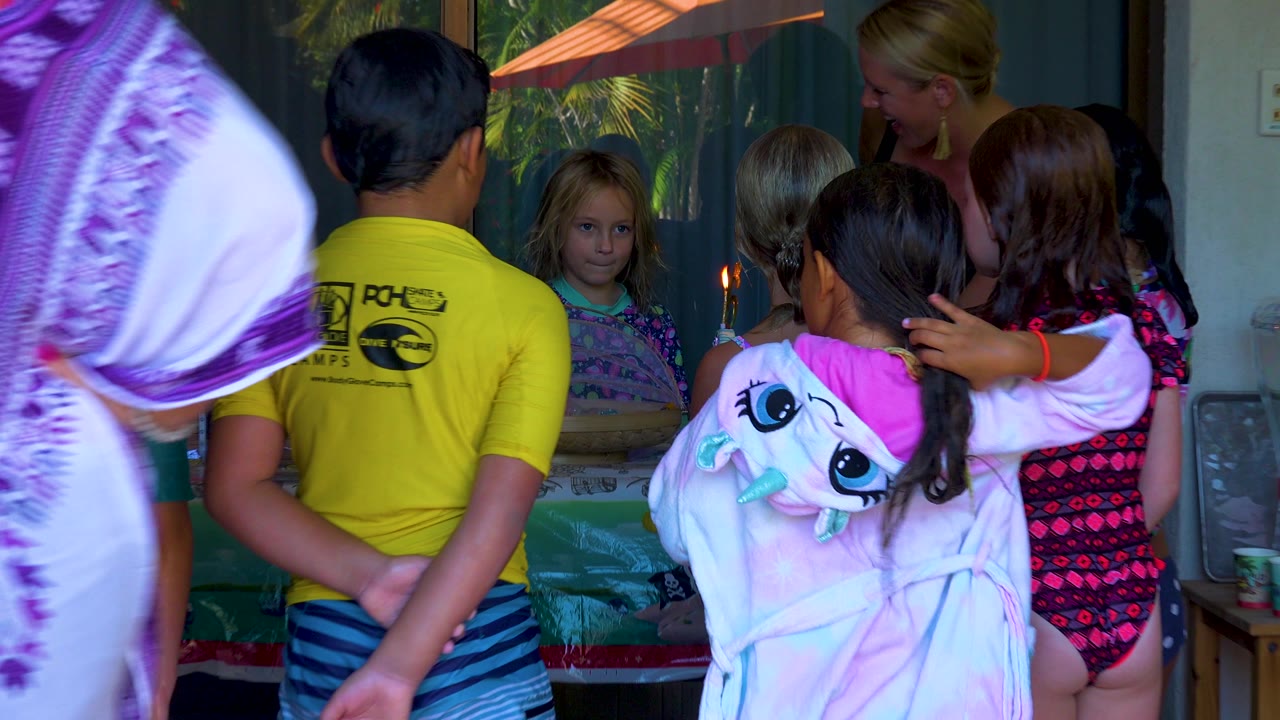 Children and an adult gather indoors around a birthday cake with a lit candle in San Jose Del Cabo, Mexico, with a boy in a yellow shirt and a girl in a pink hoodie in the foreground.