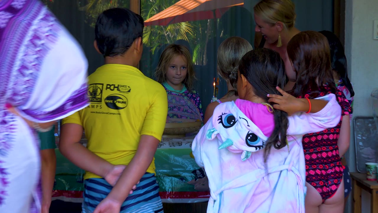 The group remains clustered around the cake in San Jose Del Cabo, Mexico, with the lit candle visible between foreground children and an adult leaning over the table.