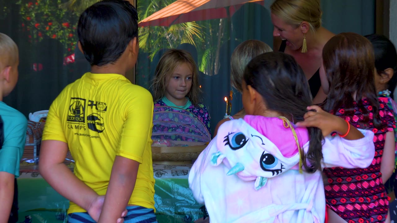 Children gather shoulder to shoulder around the cake in San Jose Del Cabo, Mexico, with the blonde girl facing the candle and foreground figures partly blocking the table.