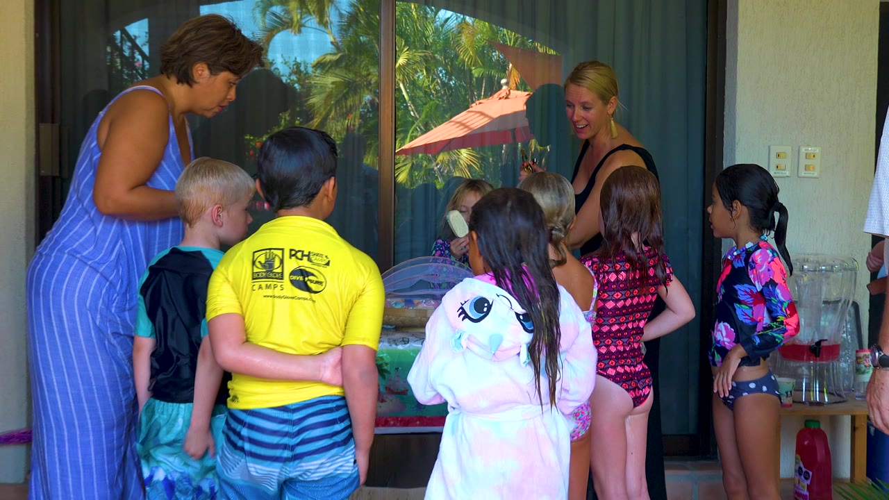 Children in swimwear and adults gather around a patio table beside reflective glass doors in San Jose del Cabo, Mexico.